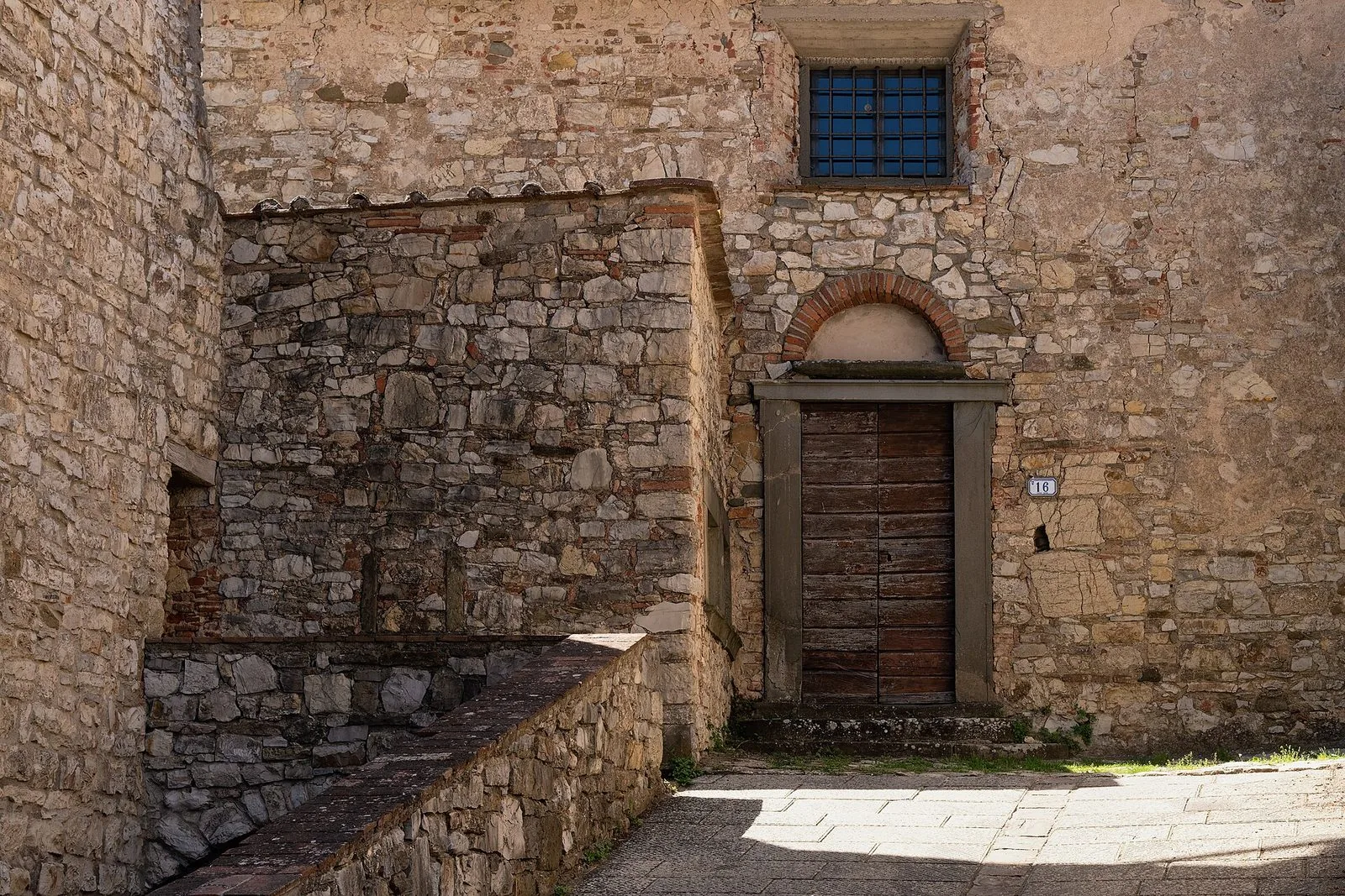 Angolo di un borgo del Chianti con muri in pietra, una porta in legno con arco in mattoni e una finestra con grata