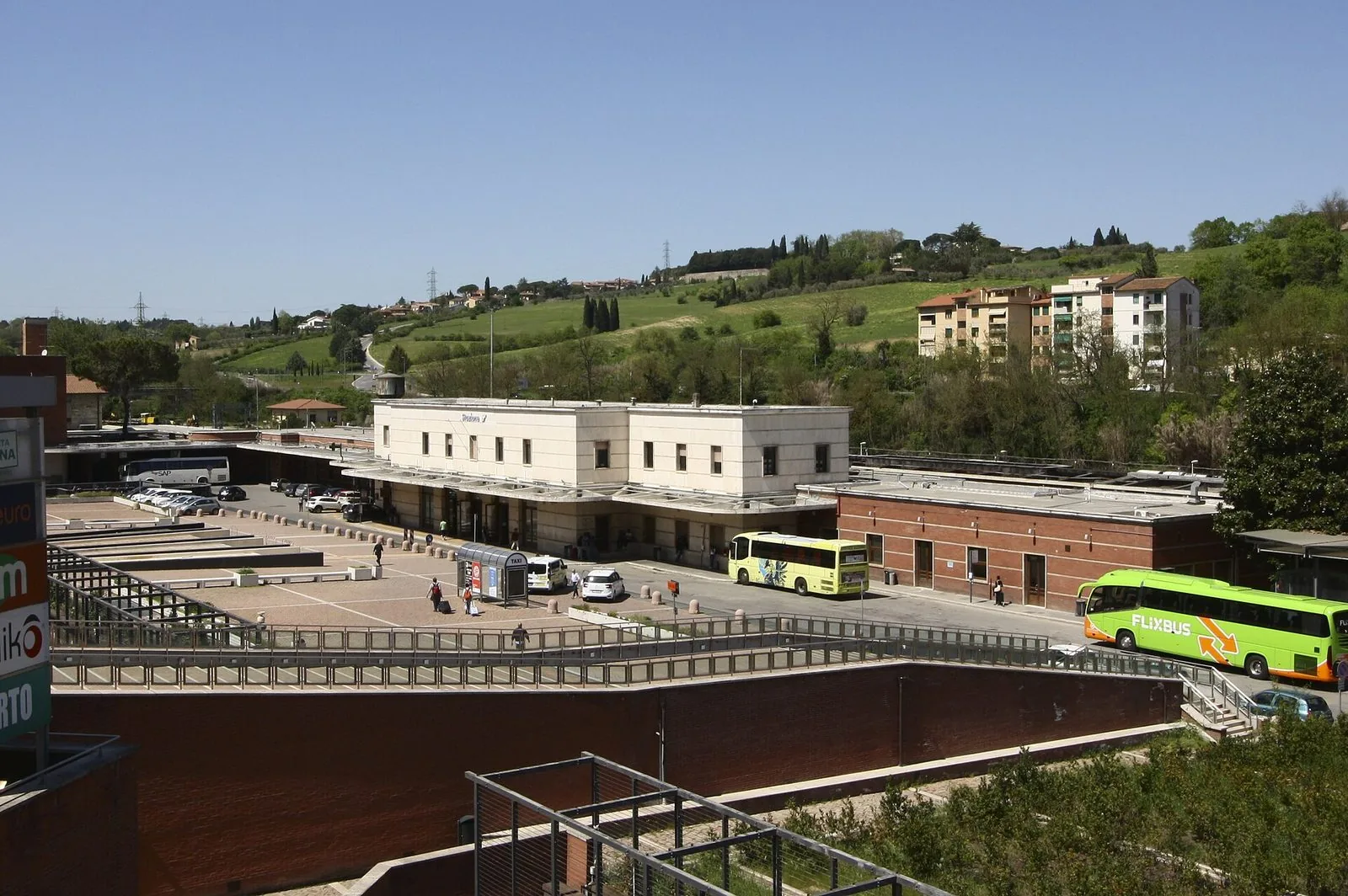Il piazzale della stazione ferroviaria di Siena con l'edificio chiaro, taxi e autobus in sosta e le colline verdi con i cipressi sullo sfondo