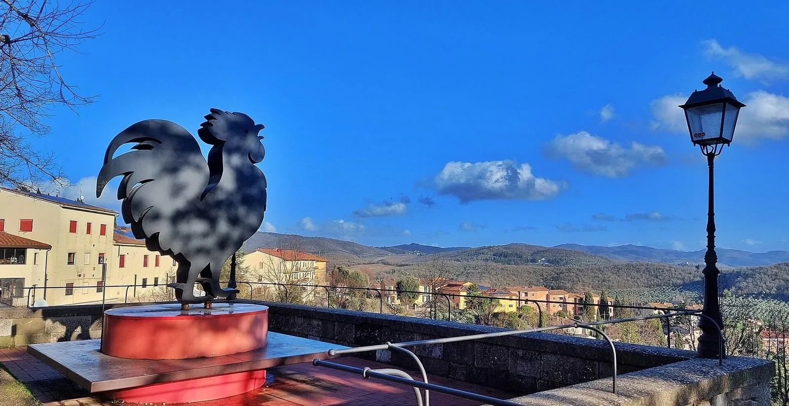 Una grande sagoma di gallo nero in metallo su un basamento rosso, su una terrazza panoramica di Radda in Chianti affacciata sulle colline boscose sotto un cielo azzurro