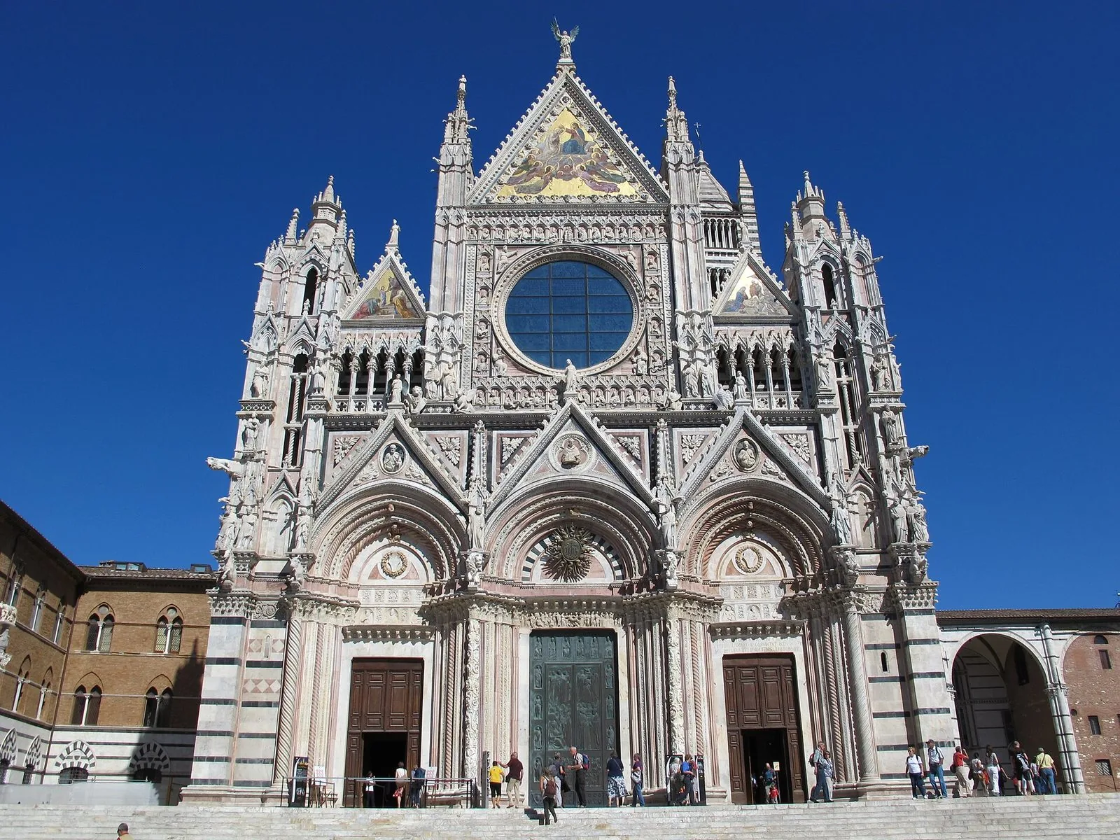 Facciata gotica del Duomo di Siena in marmo bianco e rosa con rosone, mosaici dorati e guglie sotto un cielo blu intenso, visitatori sulla scalinata